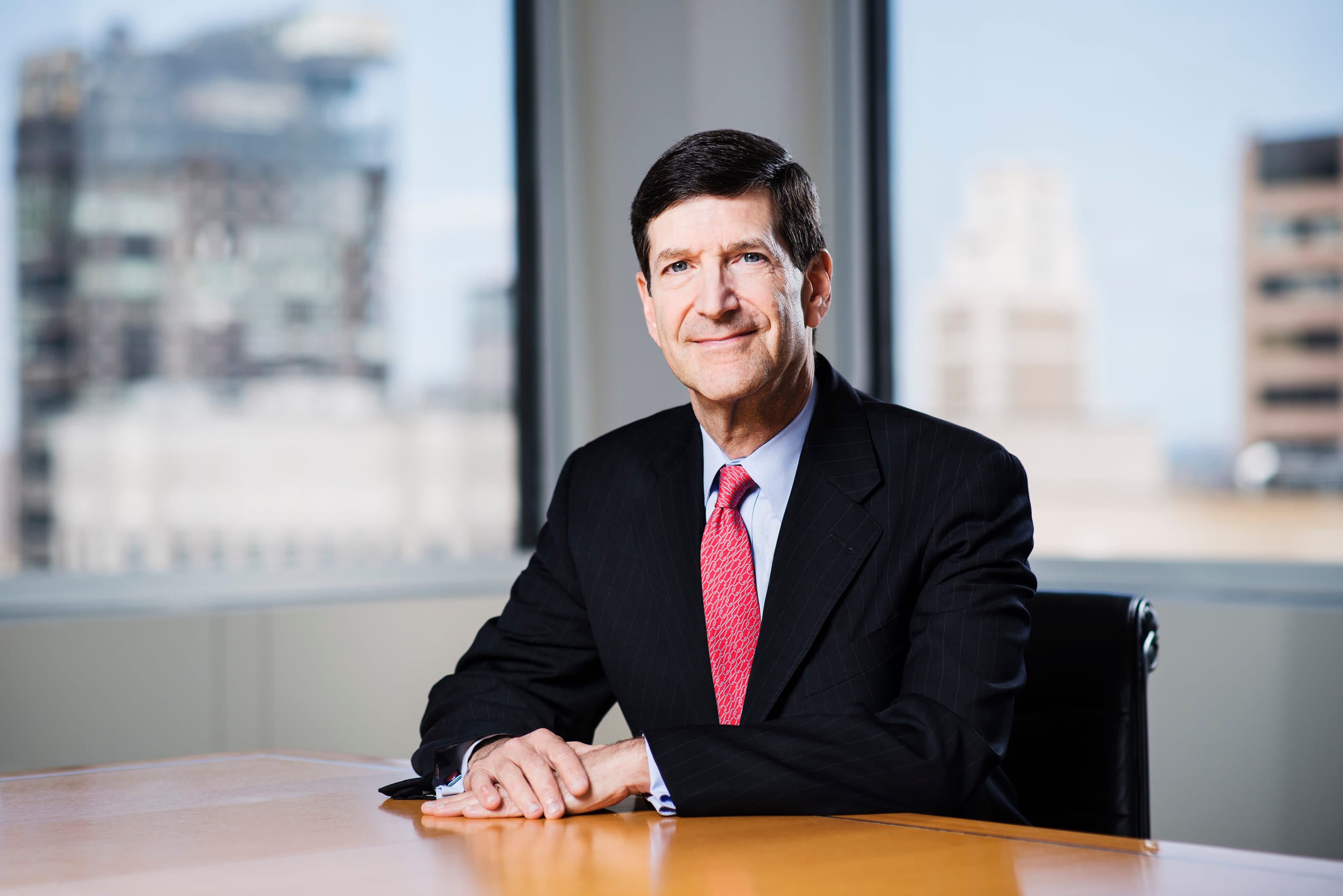 Man in a pinstripe suit and red tie sits at a table; city buildings visible.